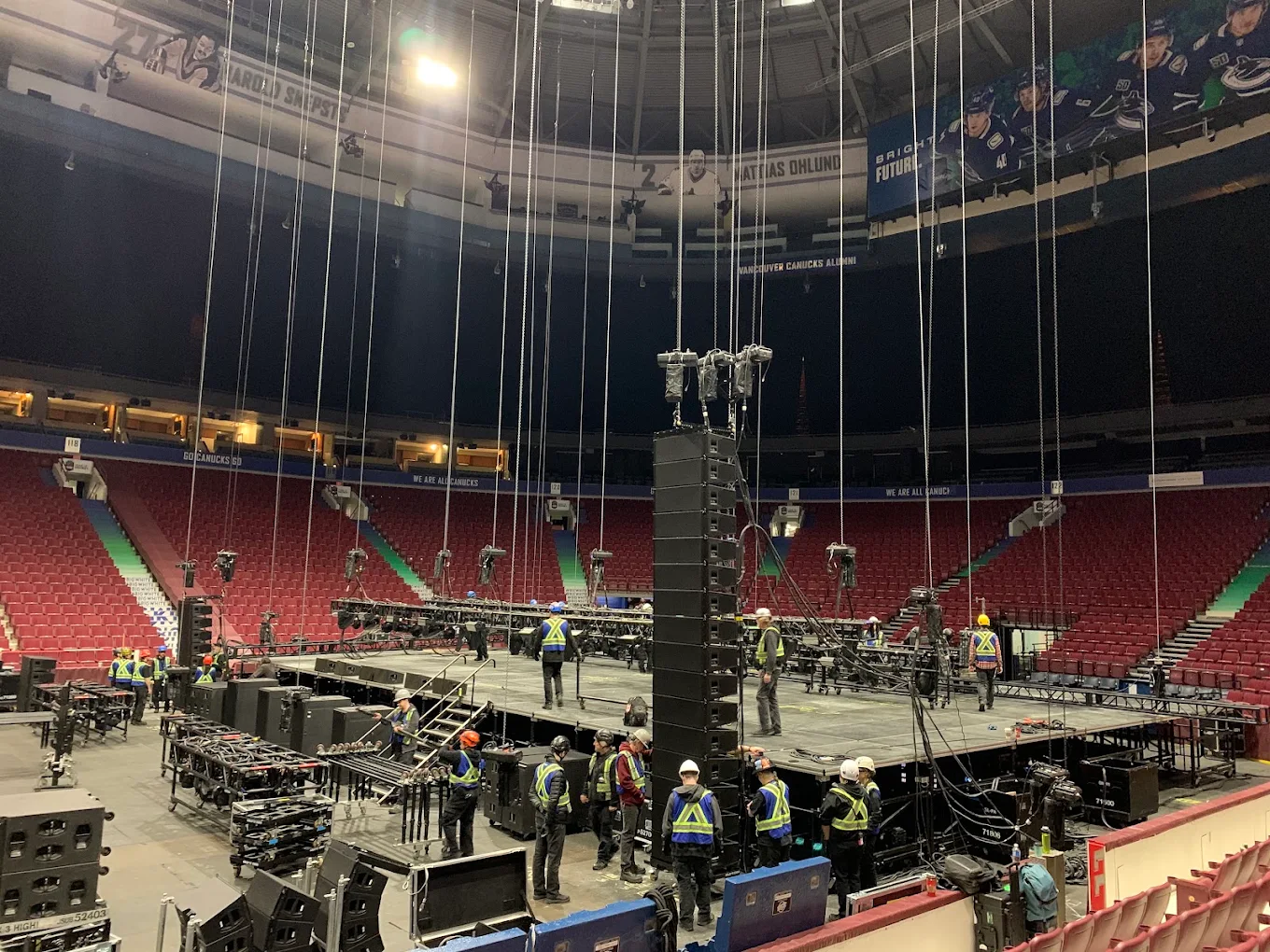A d and b line array being flown during an arena load-in, with crew rigging the stage below